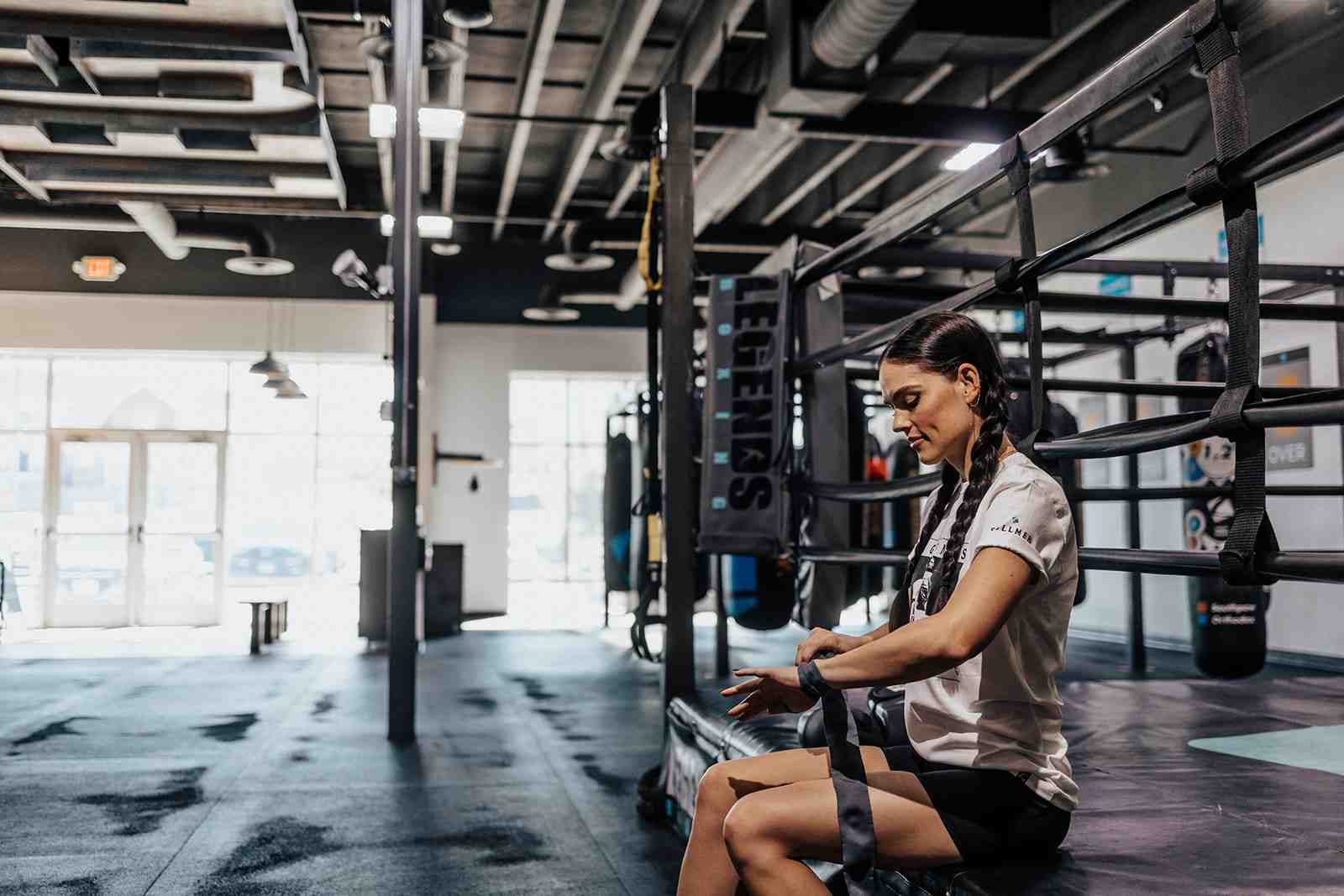 Woman with braided hair wrapping her hands while sitting on the edge of a boxing ring inside a Legends Boxing gym — natural light from windows behind her, Soneder wall rack and boxing equipment visible in the background — capturing the pre-workout ritual that every beginner should learn