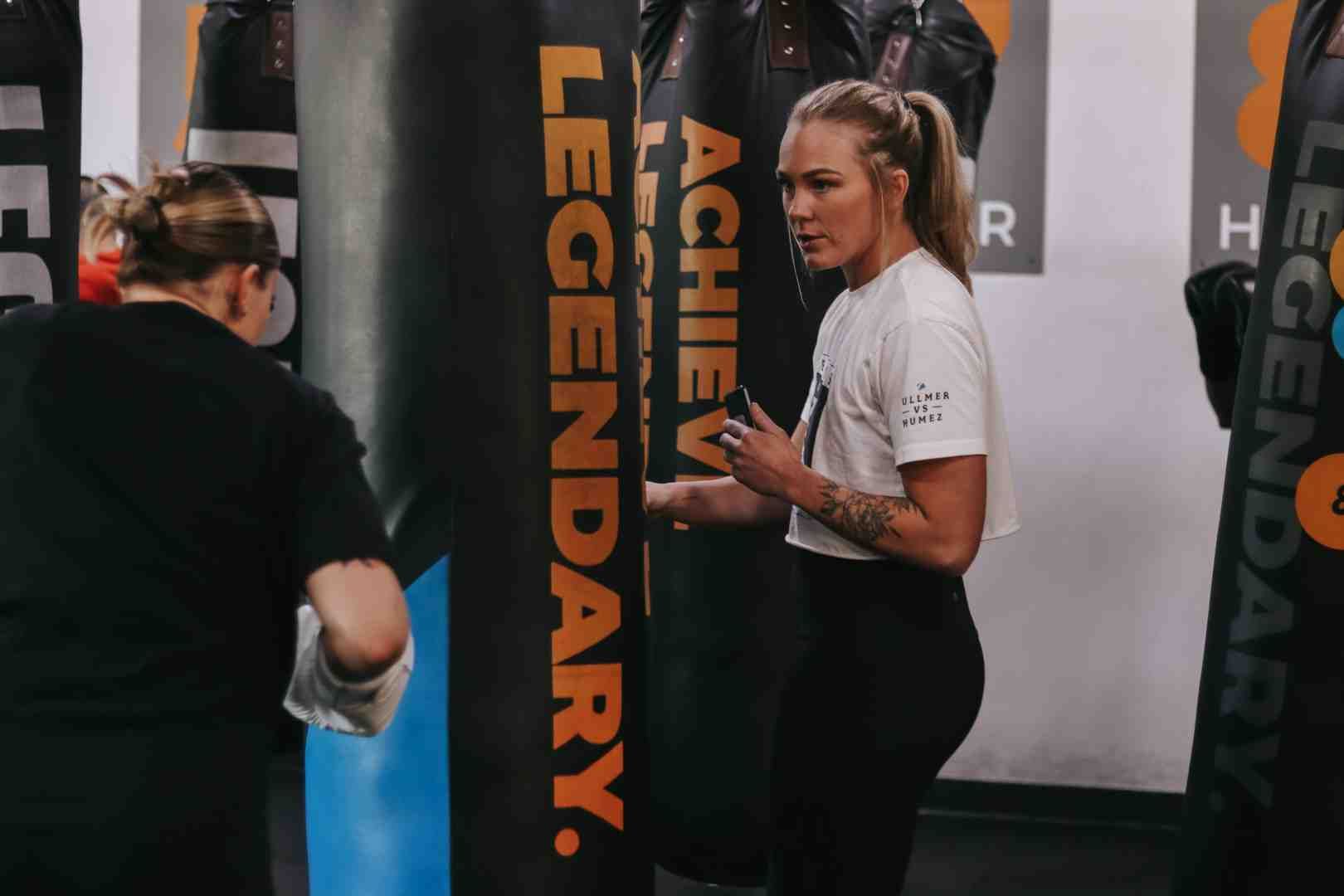 Female Legends Boxing coach with tattooed arm in white tee observing and timing a larger female member working the ACHIEVE LEGENDARY heavy bag — other members and bags visible in background — expert coaching makes the difference in beginner heavy bag circuit sessions — Legends Boxing