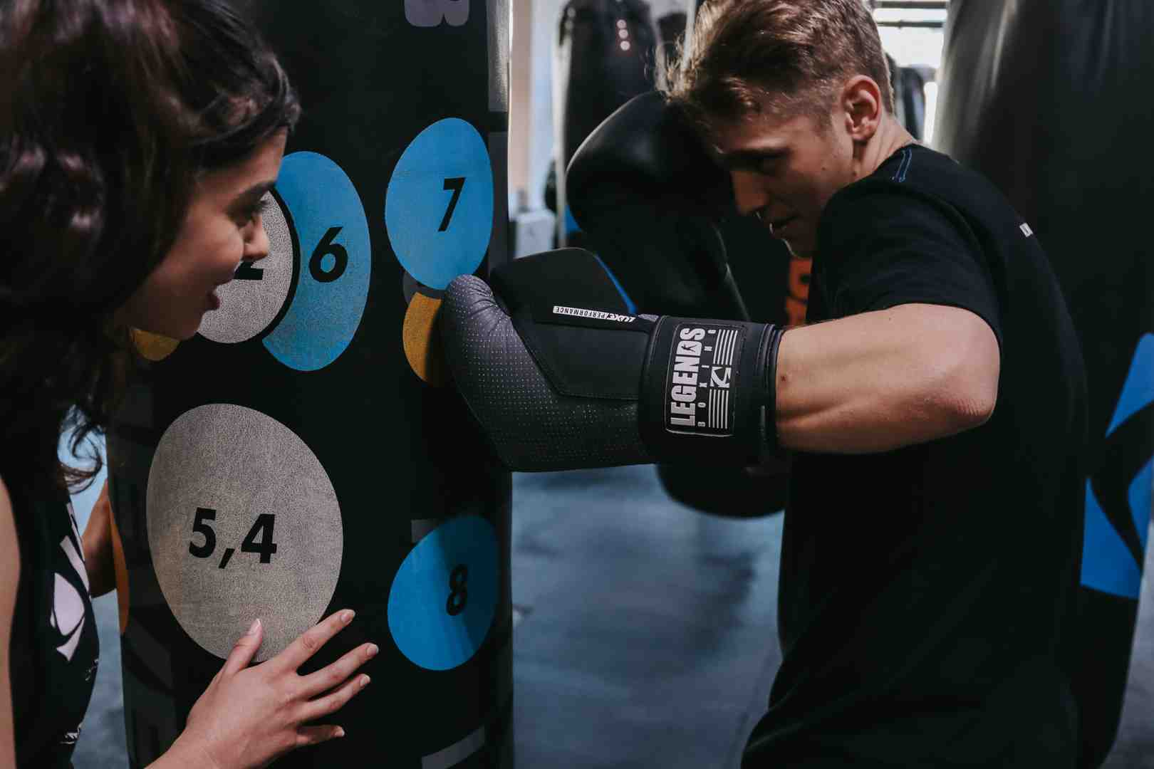 Man in Legends Boxing gloves throwing an explosive combination at a numbered heavy bag — colored target circles showing punch zones (5,4 / 6 / 7 / 8), woman observing — demonstrating the precise target focus and technique that separates effective heavy bag interval training from mindless swinging