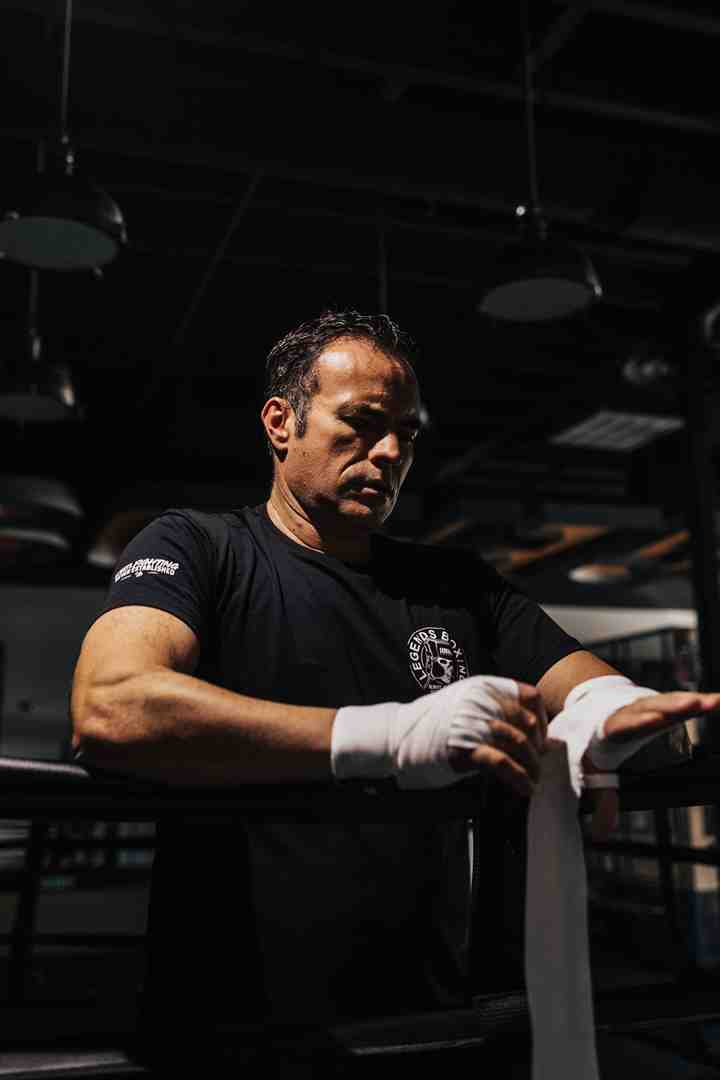 Muscular man in a Legends Boxing t-shirt carefully wrapping his hands with white boxing wraps, leaning over the ropes of a boxing ring in a dark gym — capturing the essential pre-training ritual that protects wrists and knuckles during heavy bag interval work