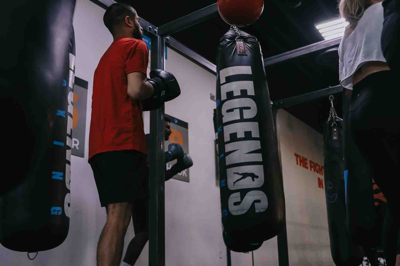 Man in red shirt resting gloves on a large "LEGENDS" branded heavy bag in a dark gym — speed bag above, another member visible nearby — authentic Legends Boxing class environment showing proper bag training setup