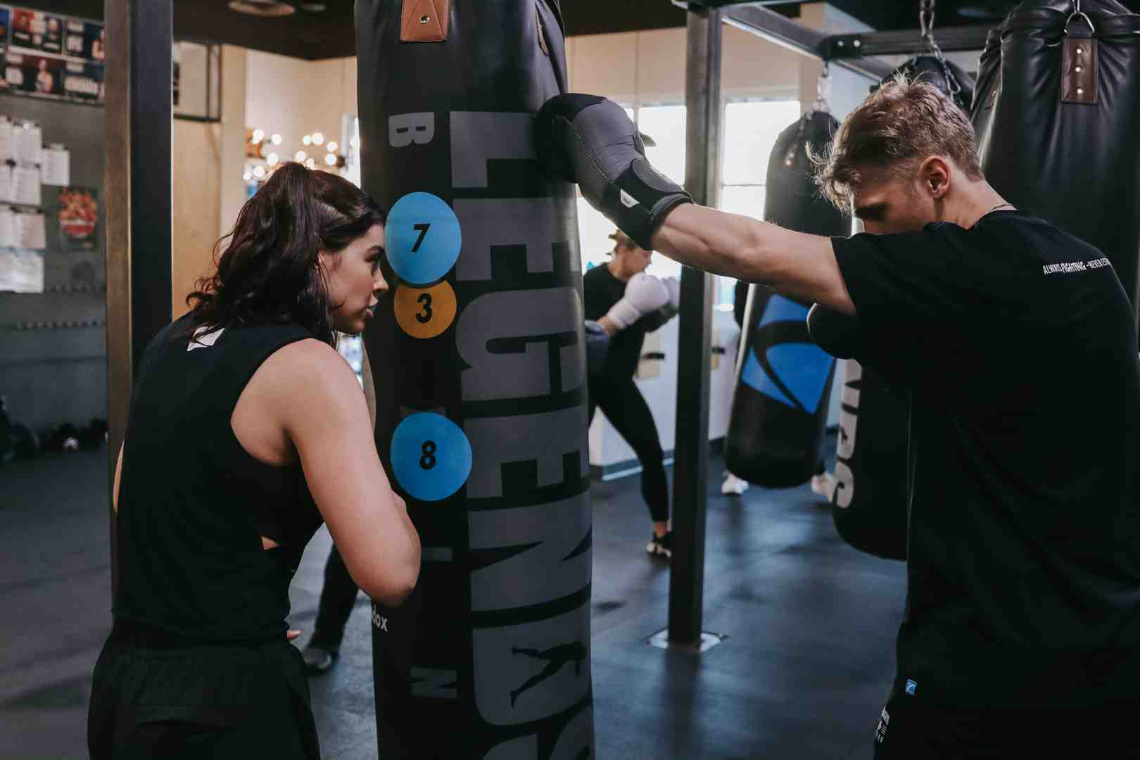 Legends Boxing class in session — woman observing partner strike numbered target circles (7, 3, 8) on a heavy bag while another member trains in the background — structured drill work building accuracy and technique — heavy bag MMA workout at Legends Boxing