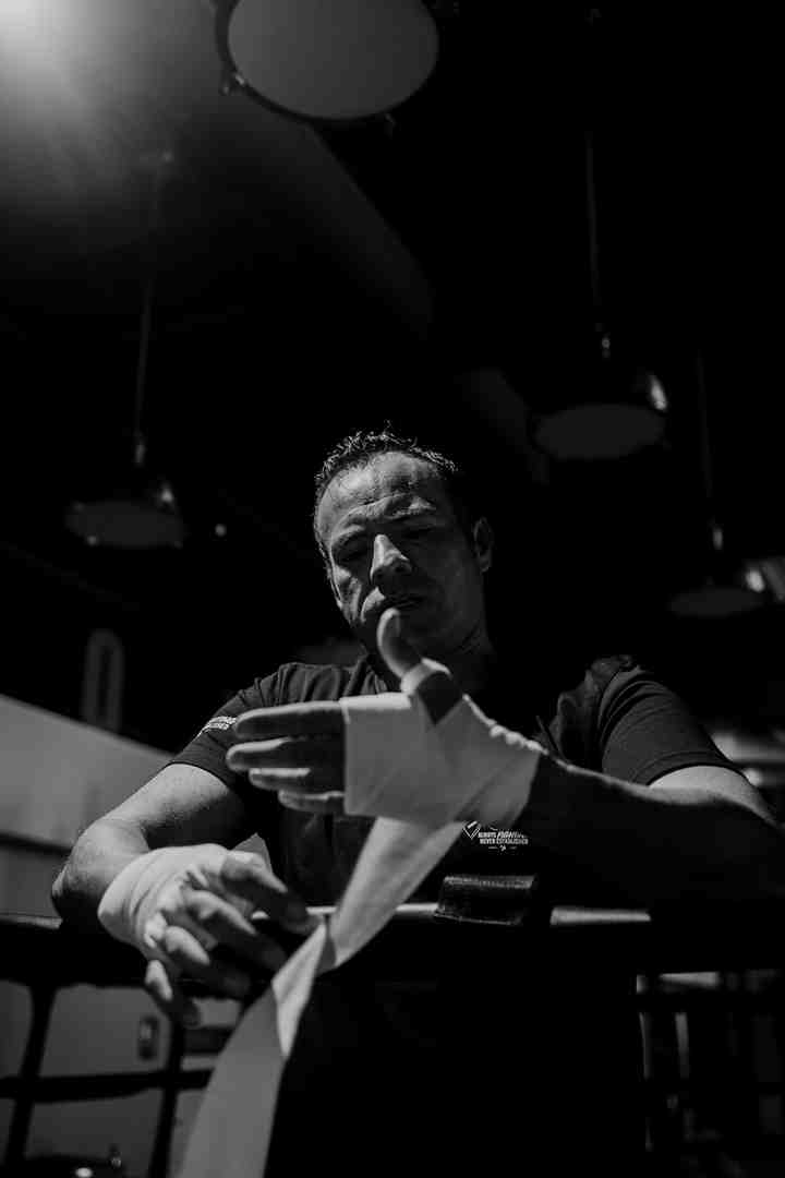 Black and white dramatic portrait of a boxer wrapping his hands while leaning on a boxing ring apron, spotlight overhead — showing the ritual of hand wrapping that every serious heavy bag practitioner treats as non-negotiable pre-training preparation