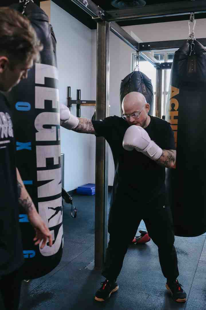 Bald man in glasses throwing a focused cross at a "LEGENDS" branded heavy bag in a bright Legends Boxing gym — proper stance and technique — another member visible in the background