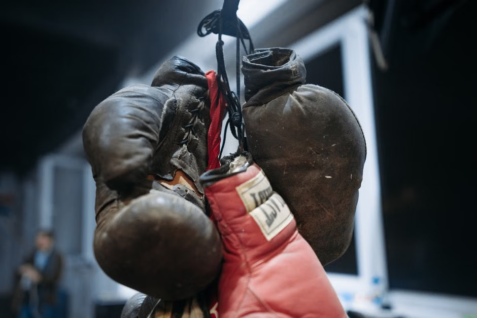 Close-up of two pairs of severely worn and deteriorated old leather boxing gloves — dark brown/black and red leather, battered seams, cracked surfaces, fraying laces — hanging together, illustrating what neglected gloves look like and when it's time for replacement