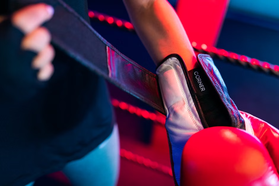 Close-up of red boxing gloves resting on ring ropes corner with 'CORNER' label visible — atmospheric red and blue gym lighting — boxing ring corner detail — Legends Boxing