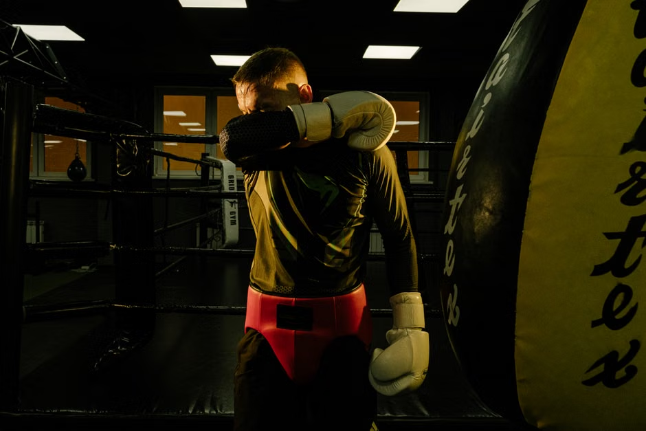 Boxer from behind wiping his gloved arm across his face in a dark gym, dramatic golden light, large heavy bag and boxing ring visible — representing the post-workout moment when glove care starts