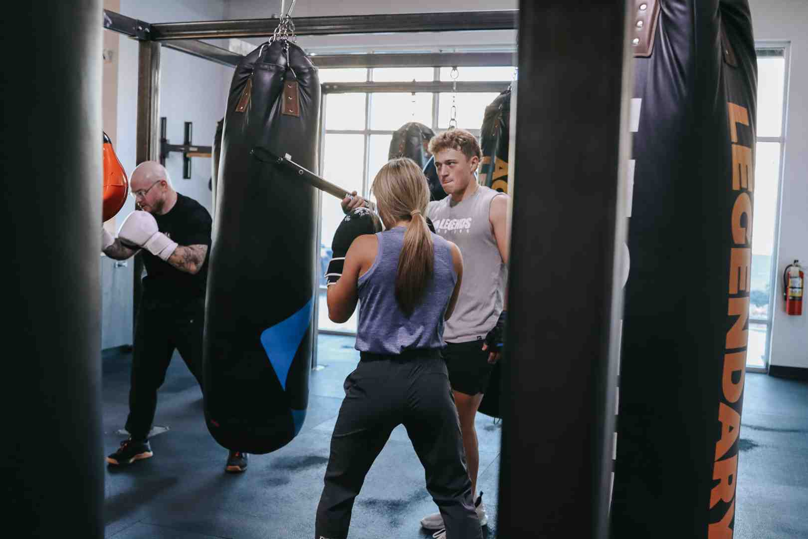 Legends Boxing coach watching a female client punch the heavy bag — 'LEGENDARY' branded bag in the foreground, natural light from large windows, second boxer training in the background — Legends Boxing Utah