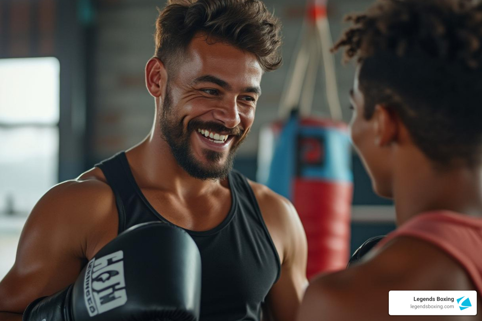Smiling male boxing coach in a gym talking with a client — heavy bags in the background, welcoming and approachable expression — Legends Boxing