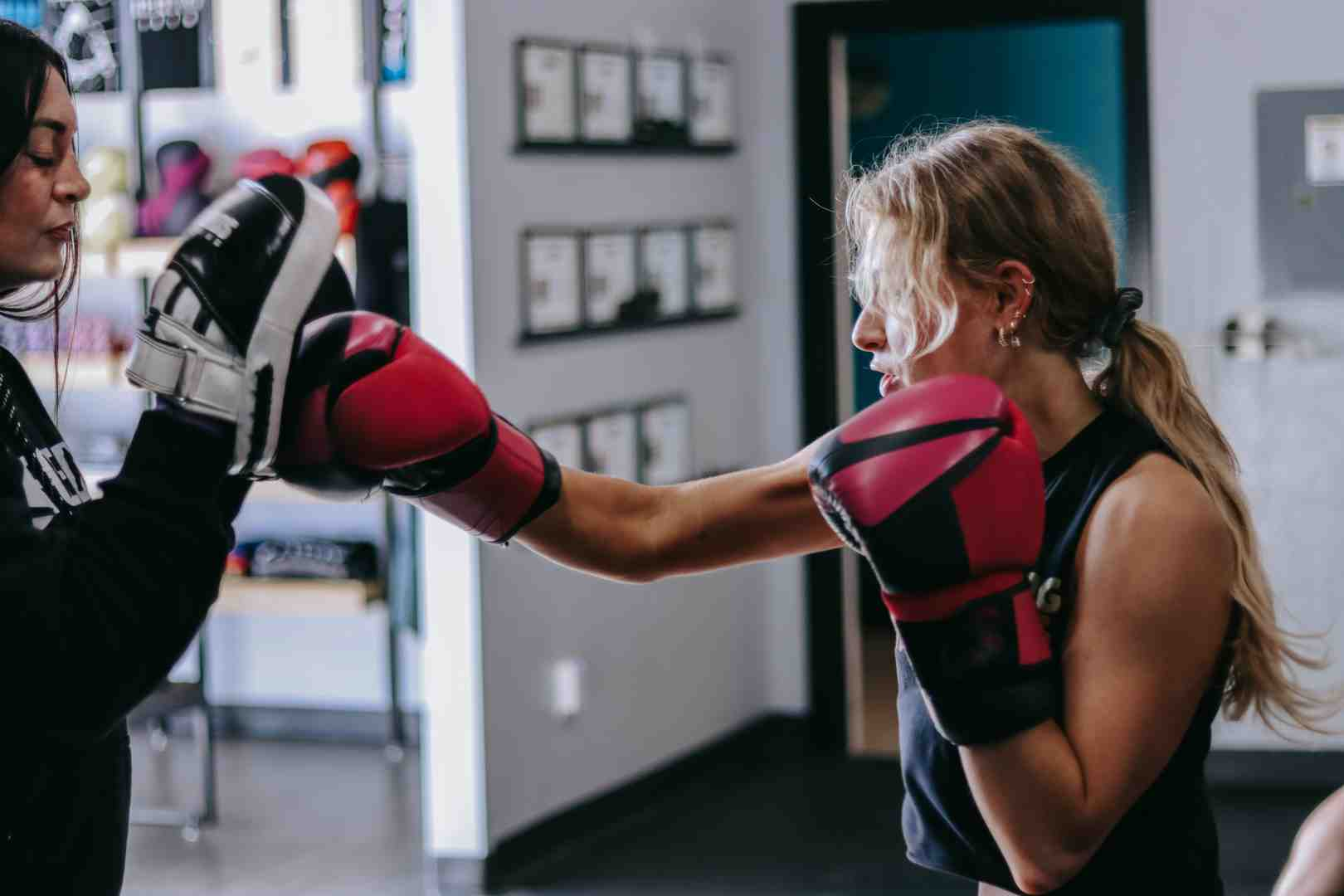 Female client throwing a jab at focus mitts held by a Legends Boxing trainer — red boxing gloves, concentrated expression, gym certificates visible on the wall in the background — Legends Boxing