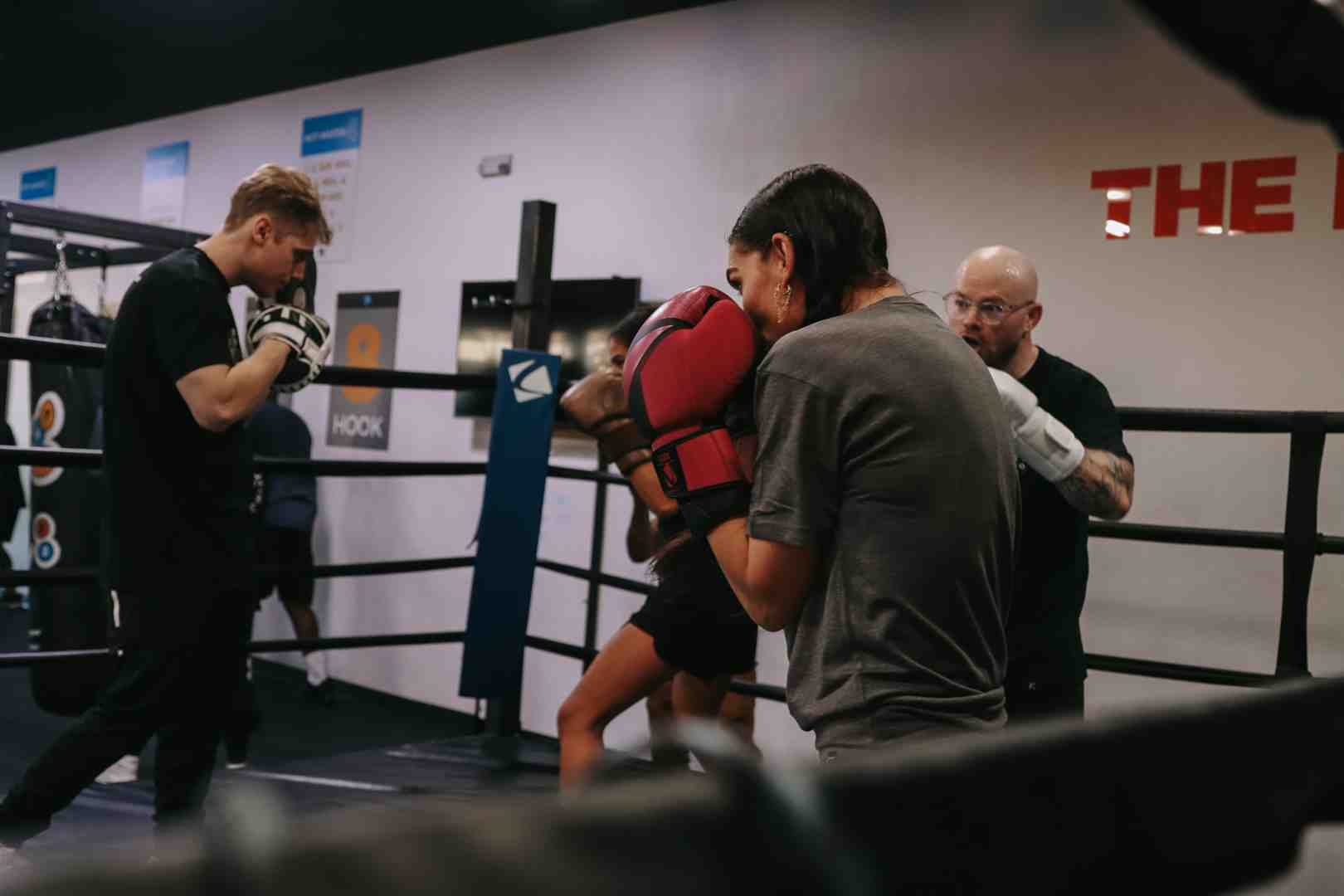 A Legends Boxing coach watches closely as a female student in red boxing gloves exchanges technique with a training partner inside a boxing ring — "HOOK" station and Legends Boxing training targets visible on the wall — showing how the Soviet emphasis on technical drilling and supervised repetition lives on in modern Legends Boxing classes