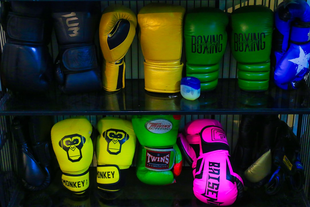 Two shelves packed with colorful boxing gloves in a gym — top shelf has navy, yellow, green, and blue gloves; bottom shelf has neon yellow Monkey brand, neon green Twins Special, and hot pink Brise gloves — a wide variety of boxing glove options — Legends Boxing